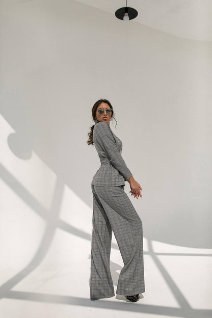 Woman wearing a gray checkered suit standing against a white wall with shadows.