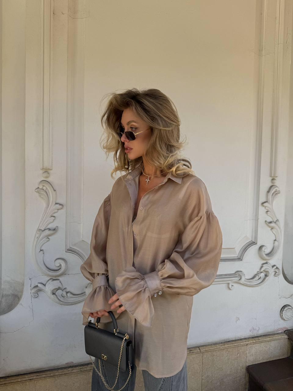 Woman wearing a sheer beige blouse with a decorative wall in the background