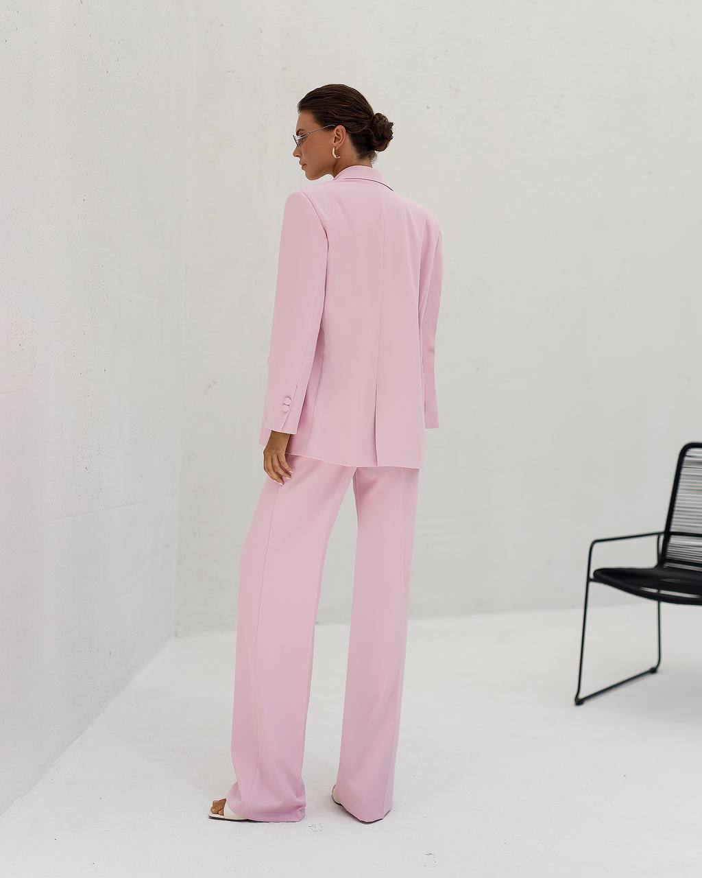 Person wearing a pink suit standing in a minimalistic setting with a black chair.