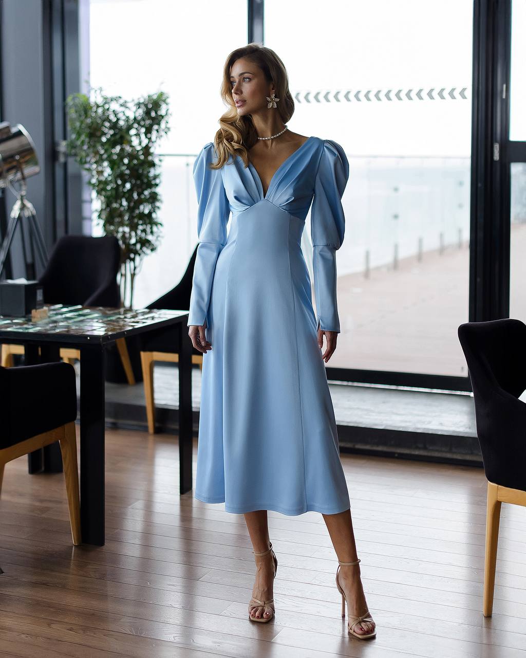 Woman in a light blue dress standing in a modern interior setting with large windows.
