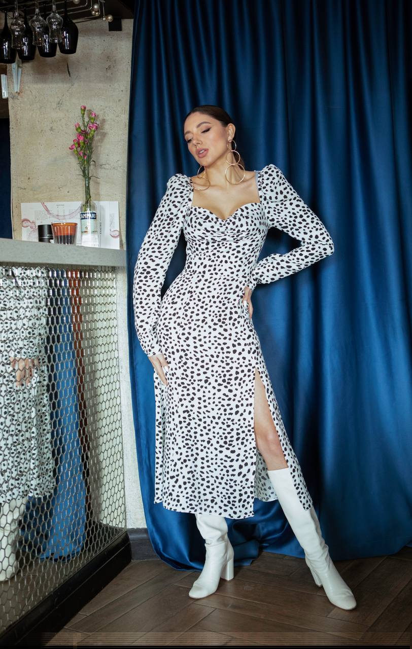 Woman in a black and white patterned dress with white knee-high boots standing in front of a blue curtain.