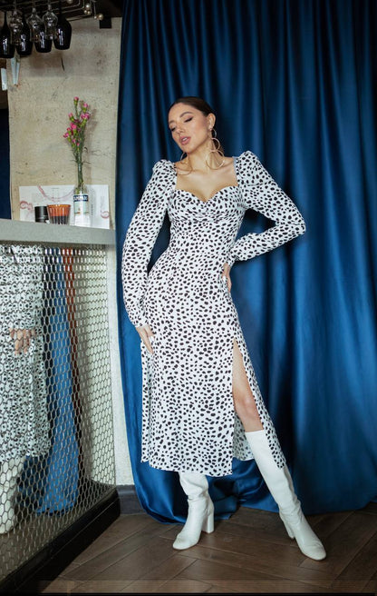 Woman in a black and white patterned dress with white knee-high boots standing in front of a blue curtain.