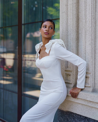 Woman in a white dress leaning against a building wall.
