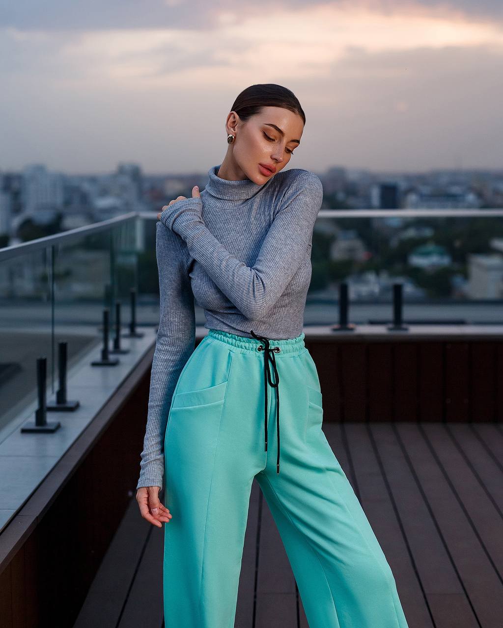 Woman in gray sweater and teal pants standing on a rooftop with cityscape background