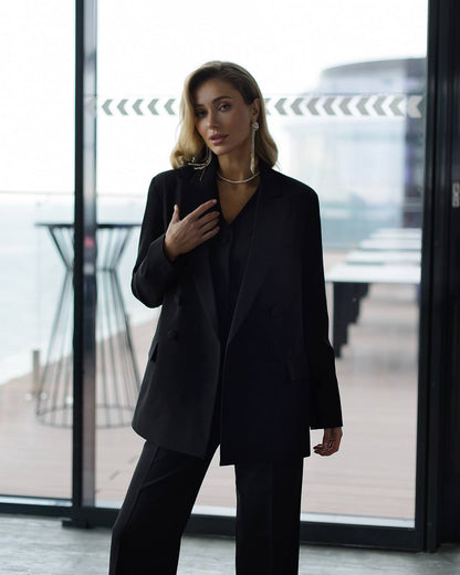 Woman in a black suit standing in an indoor setting with large windows.