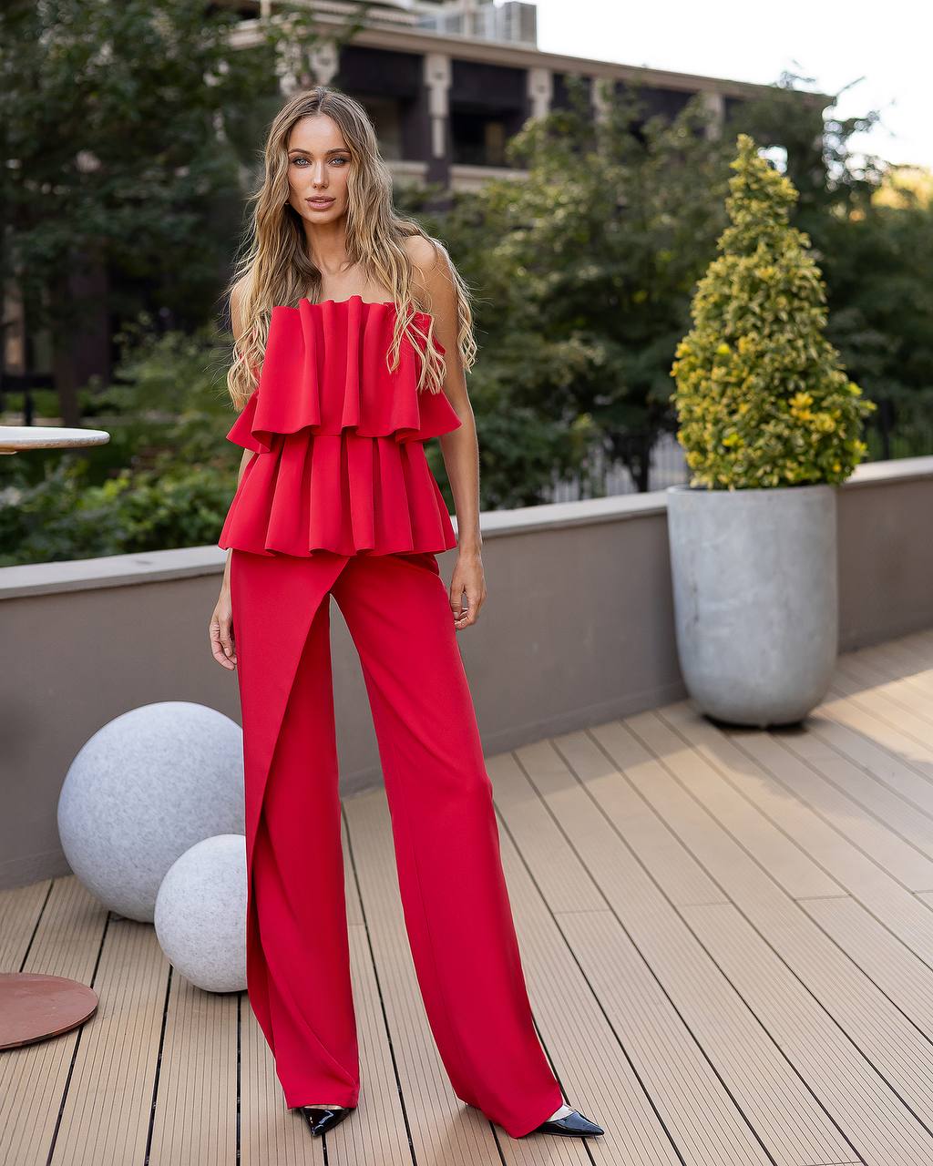 A woman modeling a bright red off-the-shoulder ruffled top and matching wide-leg trousers on a wooden deck with potted plants in the background.