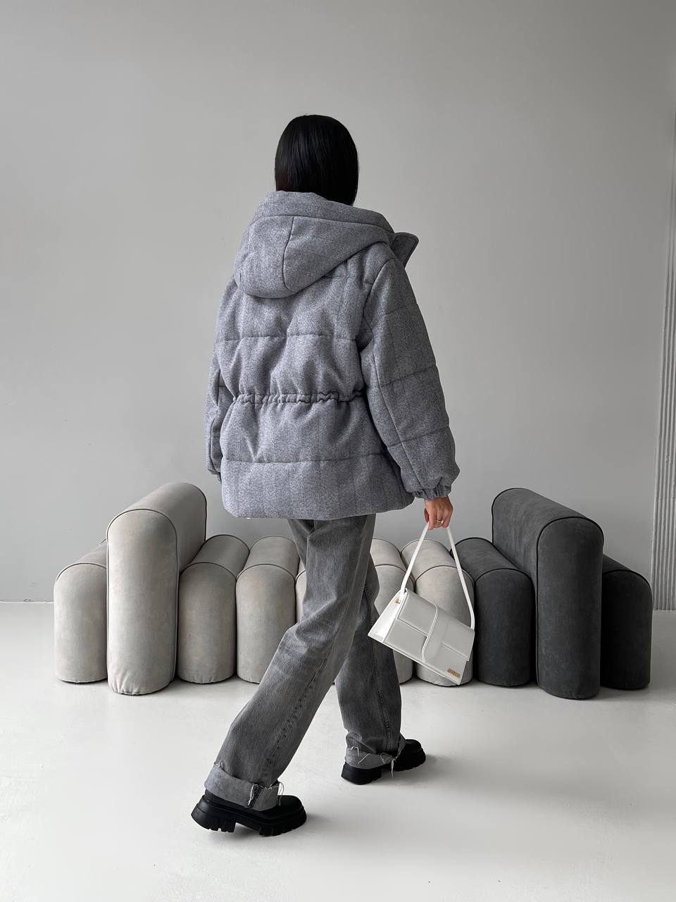 Person wearing a gray puffer jacket and pants holding a white bag, standing in front of modern gray furniture.