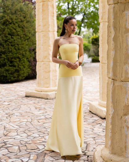 Woman in a strapless yellow and white gown standing in an outdoor setting with stone columns and greenery.