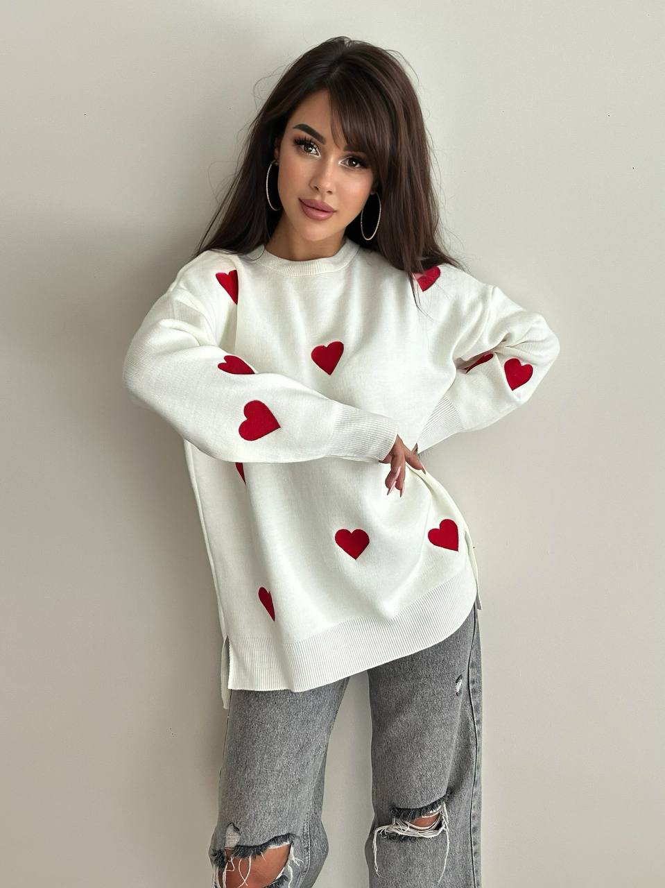 Woman wearing a white sweatshirt with red heart patterns and gray ripped jeans against a plain background