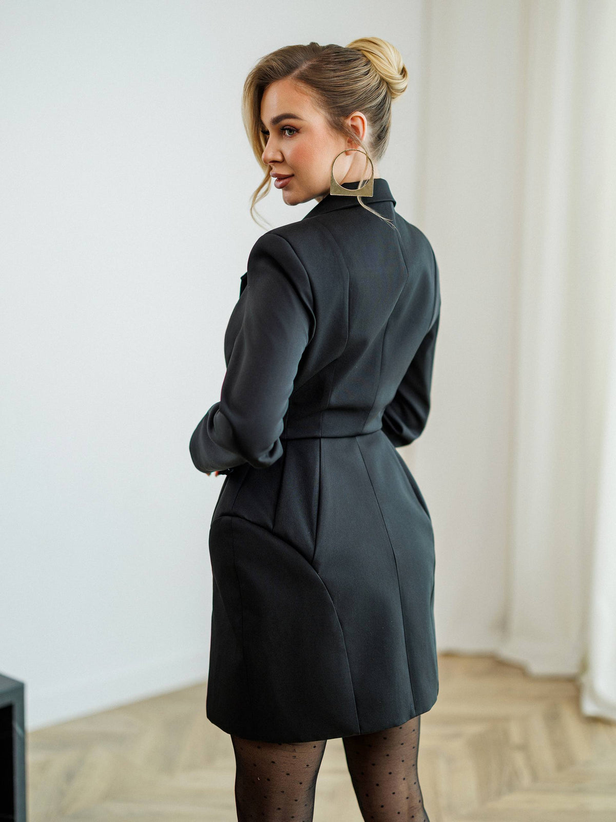 A woman wearing a dark charcoal gray wrap-style mini dress with structured shoulders and a defined waist, shown from the back in a bright indoor setting with light wood flooring and white walls.