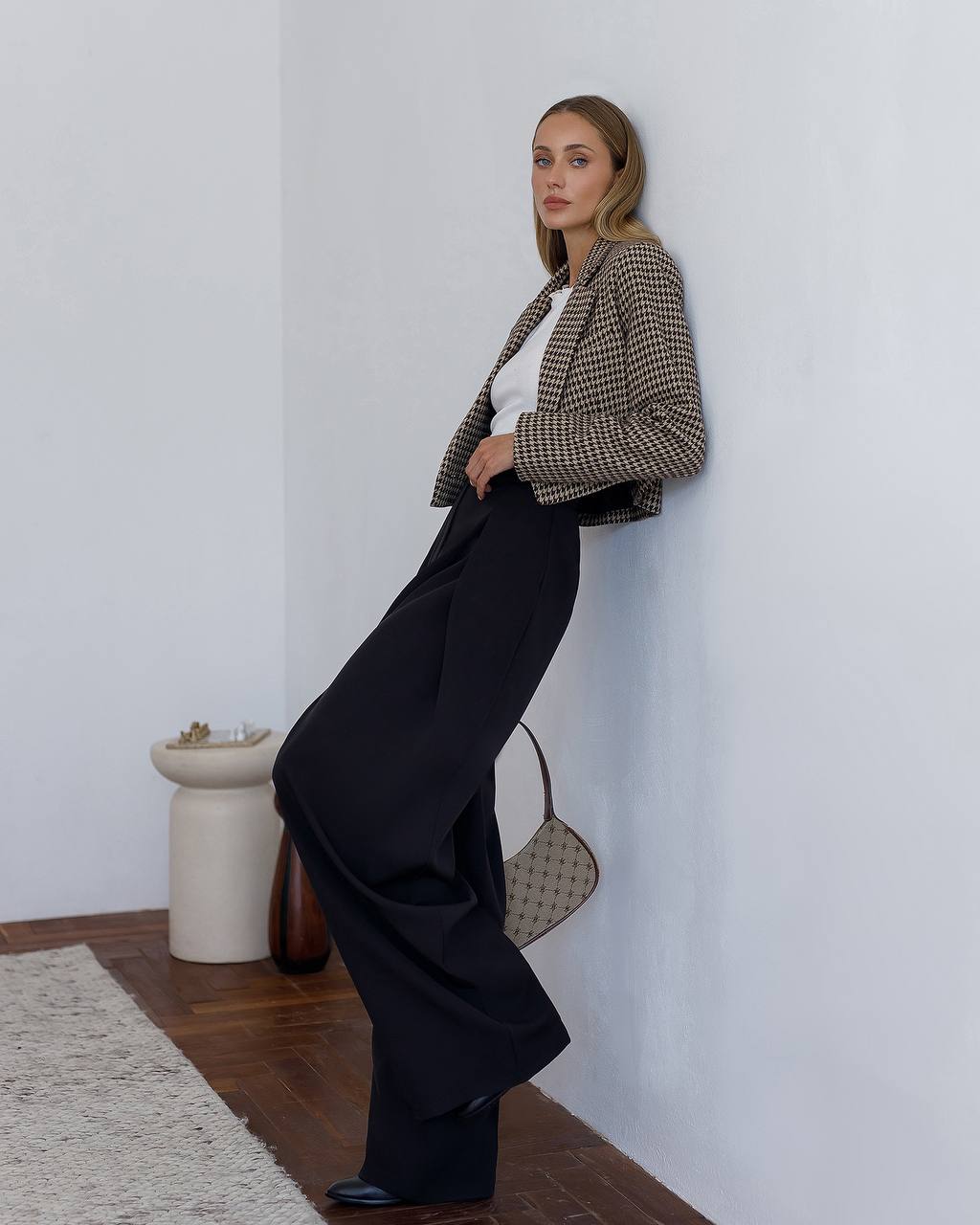 Woman in a checkered blazer and black pants leaning against a white wall.