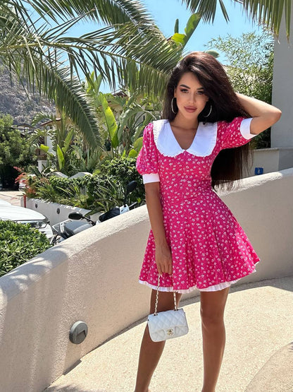Woman in a pink dress with white collar and trim standing outdoors with palm trees and buildings in the background.