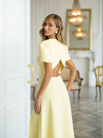 A woman wearing a pale yellow evening gown with puff sleeves and a dramatic open back, photographed in an elegant white interior room with chandeliers and a fireplace.
