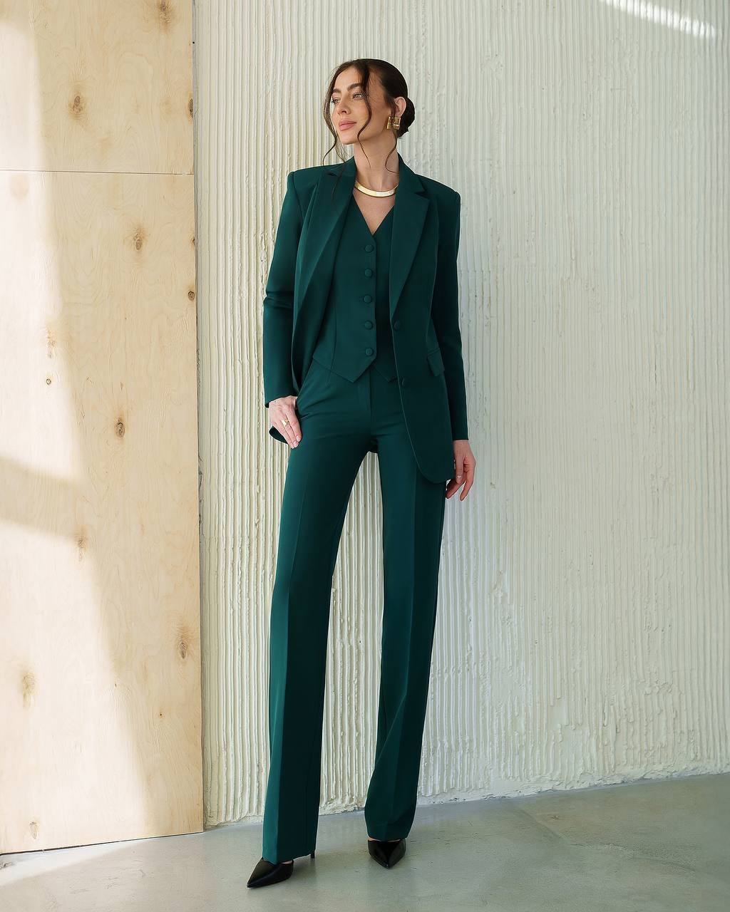 Woman wearing a dark green suit standing against a light-colored wall.