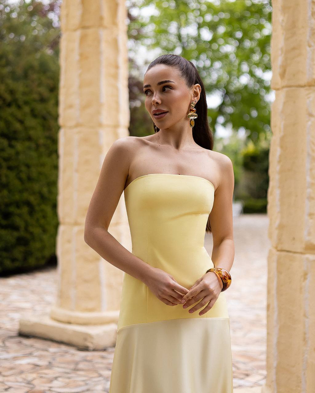 Woman in a yellow strapless dress standing outdoors with columns and greenery in the background
