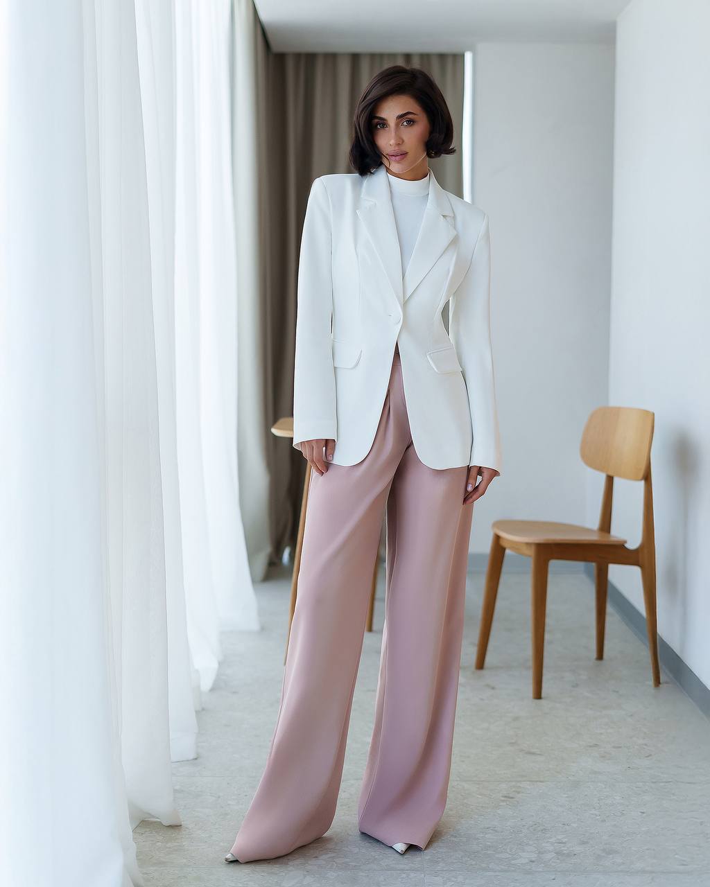 Woman wearing a white blazer and pink pants in a minimalistic room.