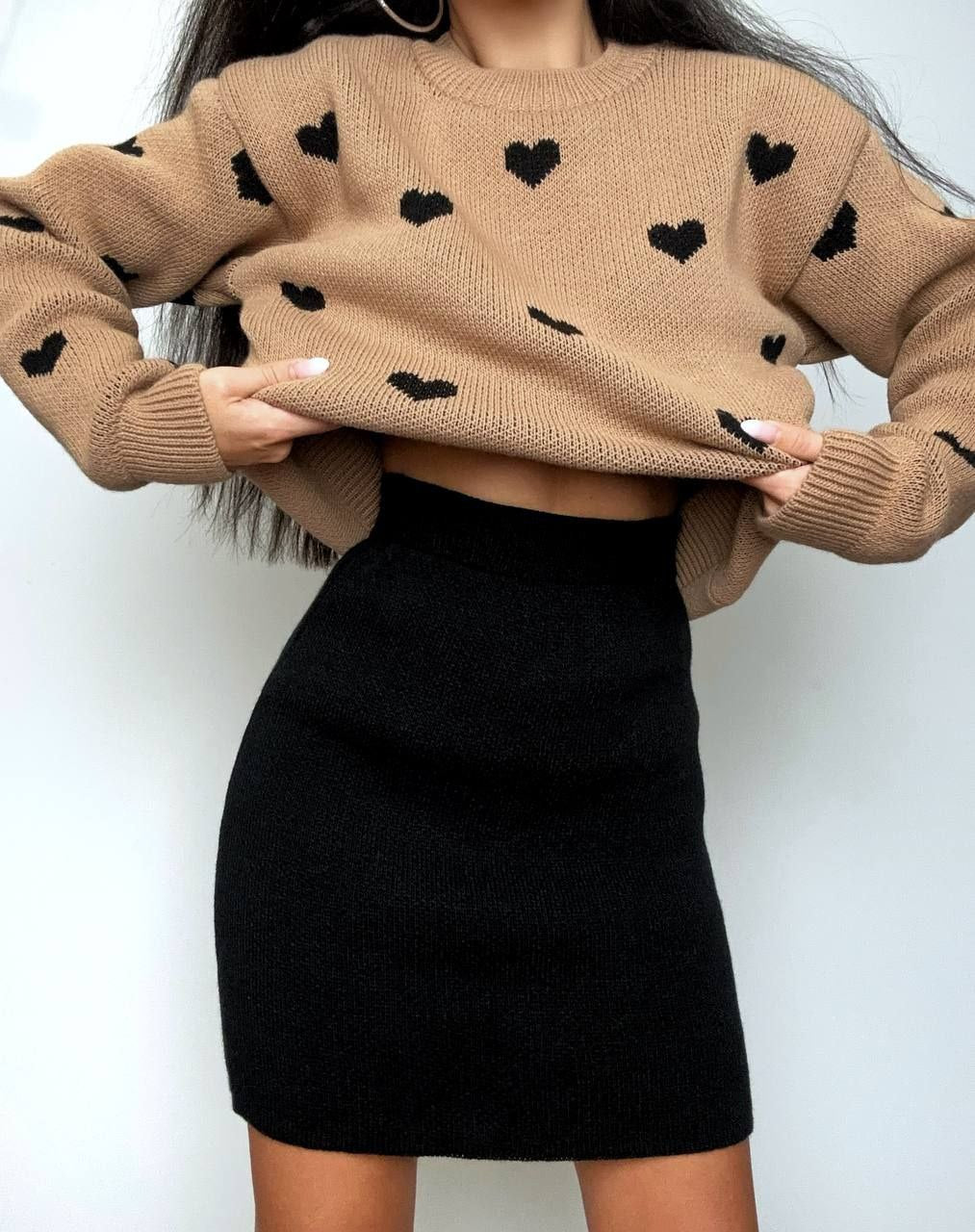 Person wearing a beige sweater with black heart patterns and a black skirt against a white background