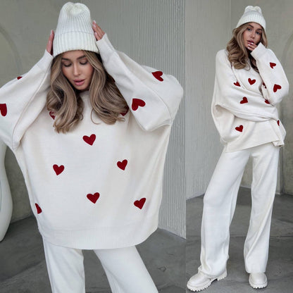 Woman wearing a white sweater with red heart patterns and white pants, standing against a neutral background.