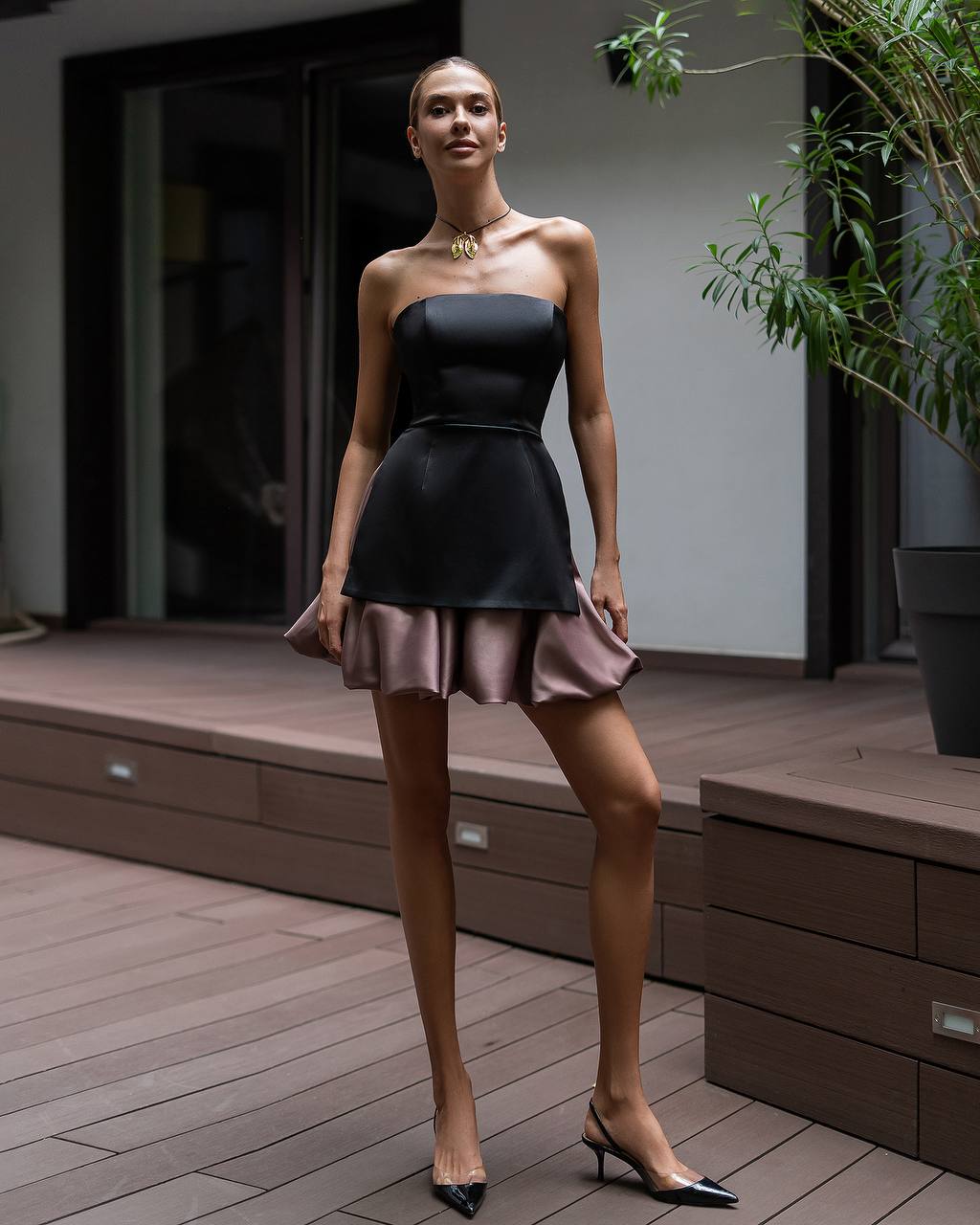 A woman models a strapless black mini dress with a flared skirt and a dusty rose tulle hem, paired with black pointed-toe heels, standing on a wooden deck.