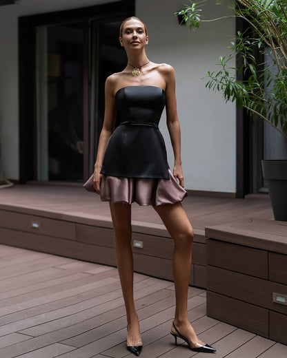 A woman models a strapless black mini dress with a flared skirt and a dusty rose tulle hem, paired with black pointed-toe heels, standing on a wooden deck.