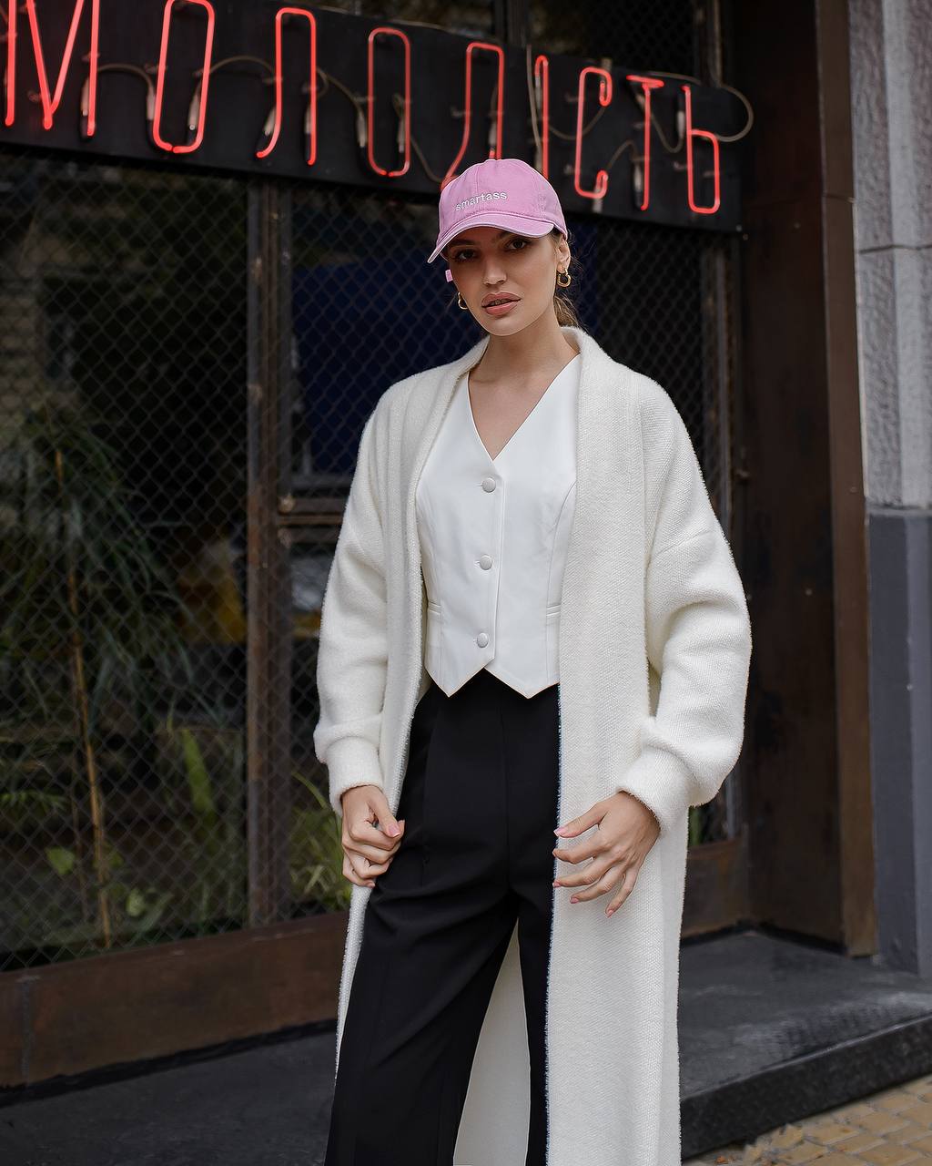 Woman wearing a white cardigan, white blouse, black pants, and pink cap in front of a neon sign.