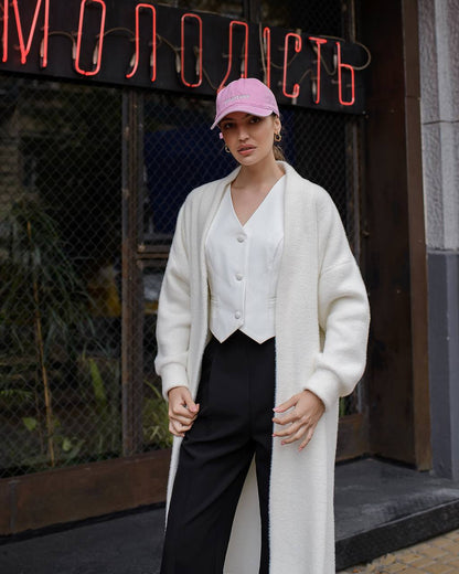 Woman wearing a white cardigan, white blouse, black pants, and pink cap in front of a neon sign.
