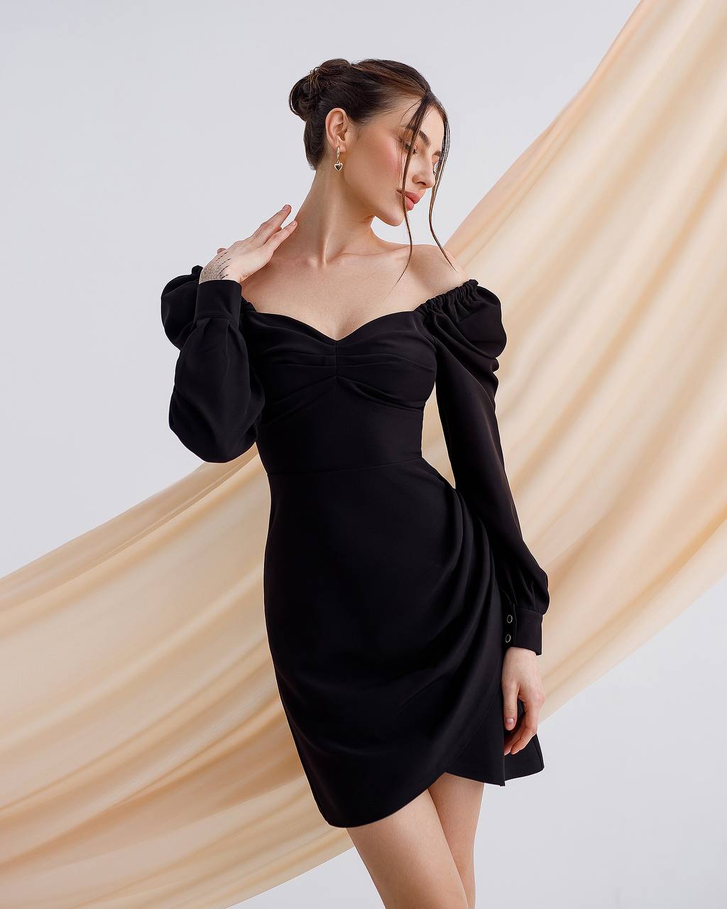 Woman wearing a black off-shoulder dress with long sleeves against a beige curtain background