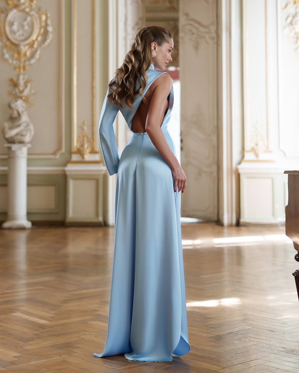 Woman in a light blue evening gown standing in an elegant room with wooden floor and decorative walls.