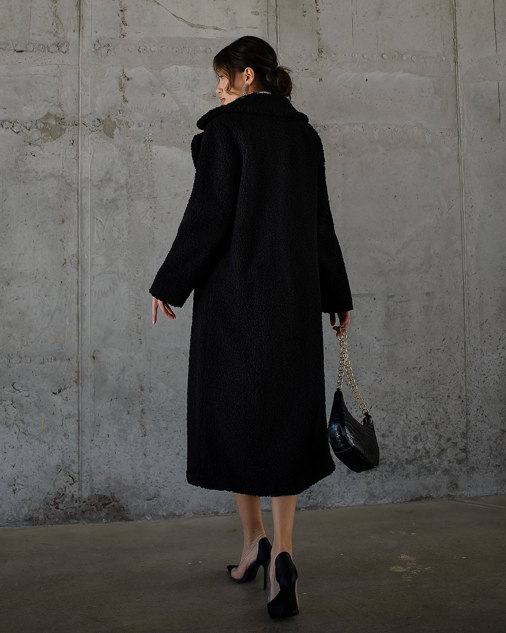 Person wearing a black coat holding a black handbag against a gray concrete wall.