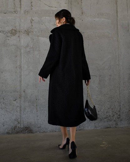 Person wearing a black coat holding a black handbag against a gray concrete wall.
