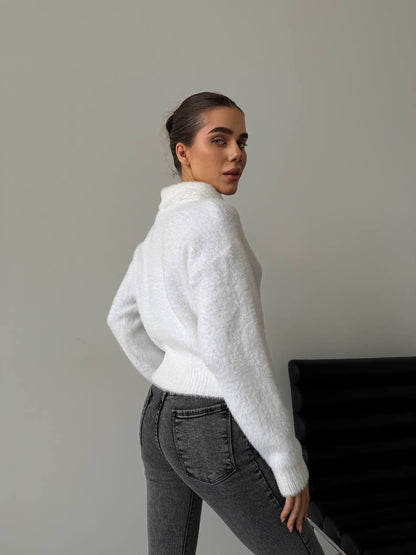 Person wearing a white turtleneck sweater and gray pants against a plain background