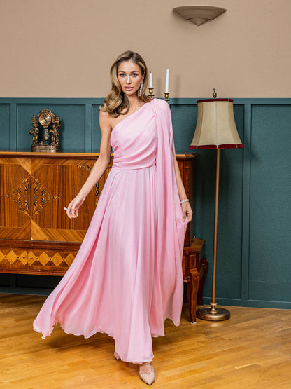 Woman in a pink one-shoulder dress standing in a room with wooden furniture and a lamp.