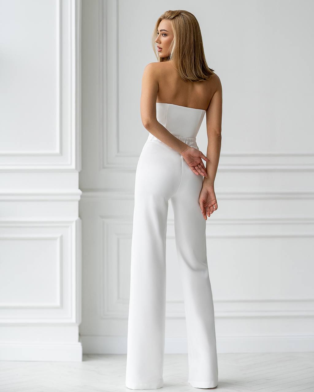Woman wearing a strapless white jumpsuit against a white wall.