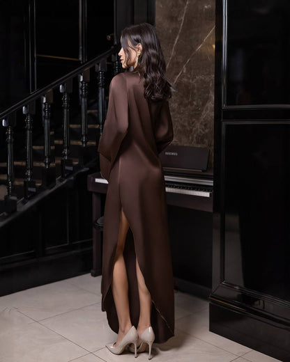 A woman stands in a luxurious interior with a staircase and piano, wearing a long, dark brown satin-like dress with a high thigh slit and draped sleeves, paired with beige high heels.
