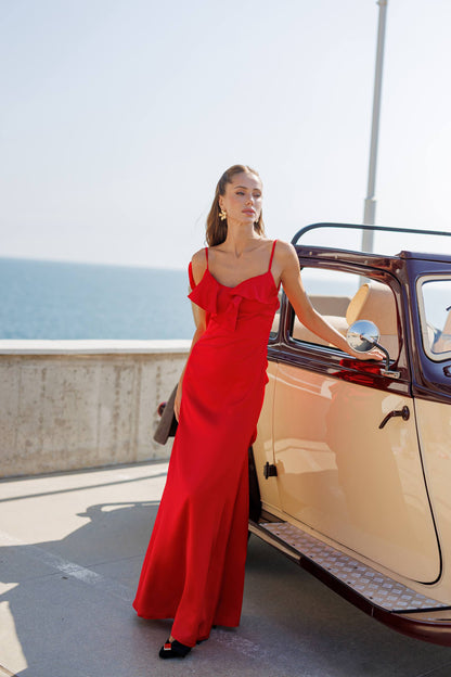 Woman in a red dress standing next to an open car by the sea.