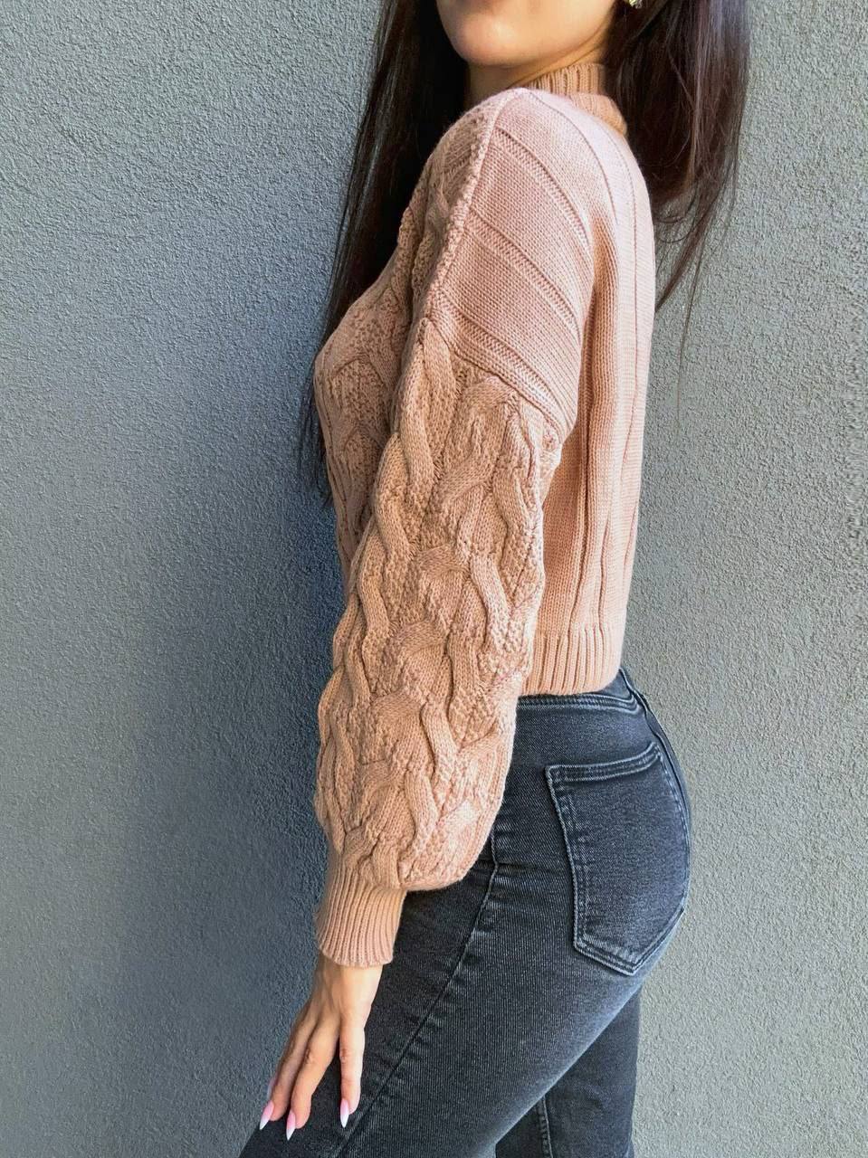 Person wearing a beige cable knit sweater and dark jeans against a gray wall