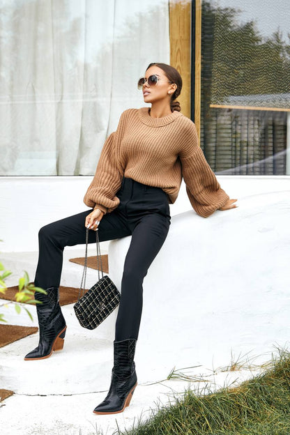 Woman in a beige sweater and black pants sitting on a white ledge outdoors.