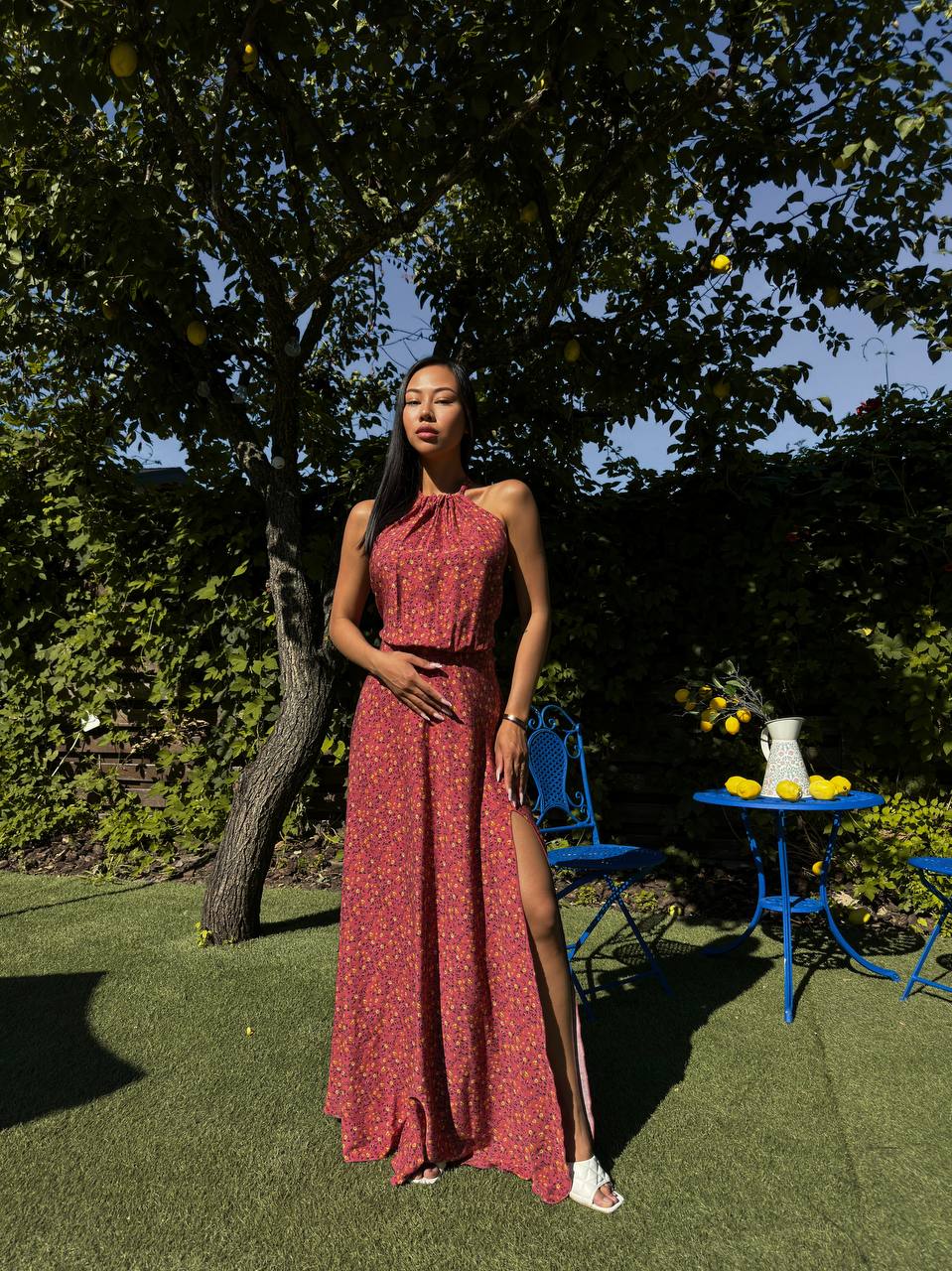 Woman in a red dress standing outdoors with greenery and a blue chair in the background
