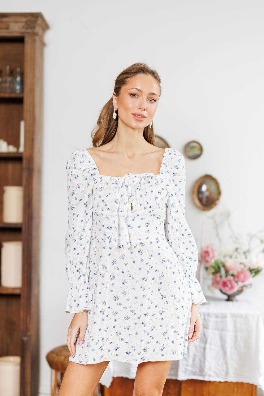 Woman wearing a white floral dress in a room with a wooden shelf and table.