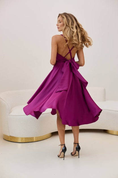 Woman in a purple dress with a back bow, standing in a minimalistic room.