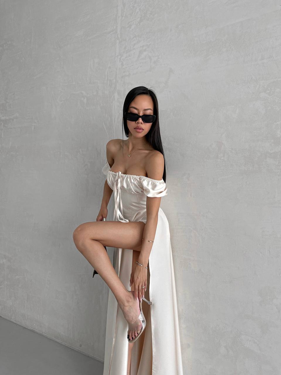 Woman in a white off-shoulder dress with a high slit, standing against a plain gray wall.