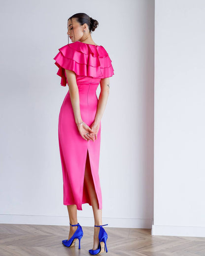 Woman wearing a bright pink dress with ruffled top and high slit, standing against a white wall.