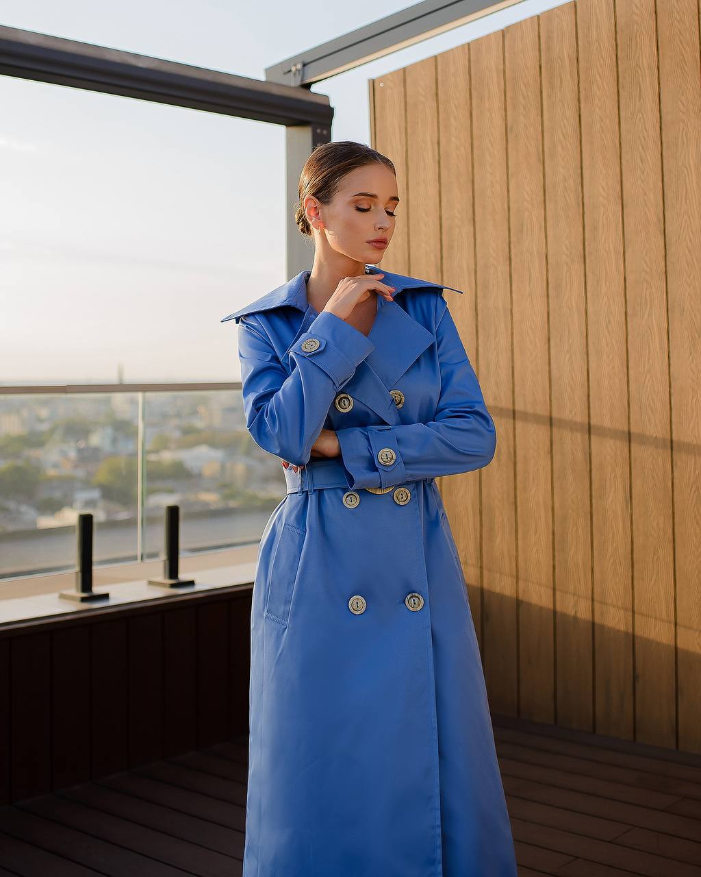 Woman wearing a blue trench coat on a balcony with a cityscape view.