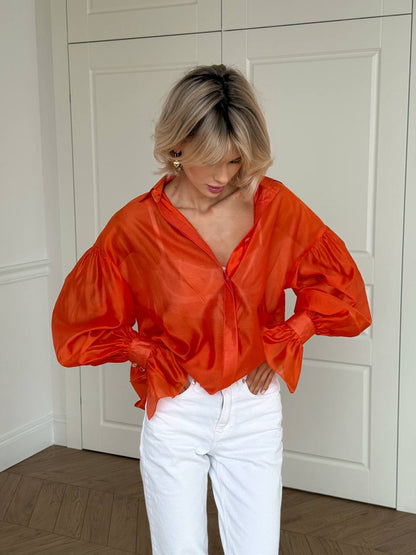 Person wearing an orange satin blouse and white pants in a room with white walls and wooden floor.