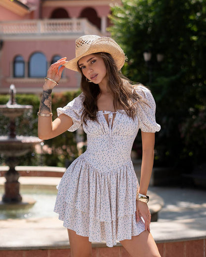 Woman in a white floral dress and straw hat standing in front of a fountain and building.