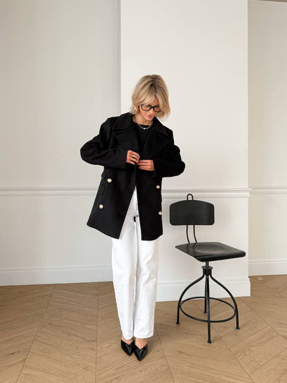 Person wearing a black coat and white pants standing in a room with a black chair.