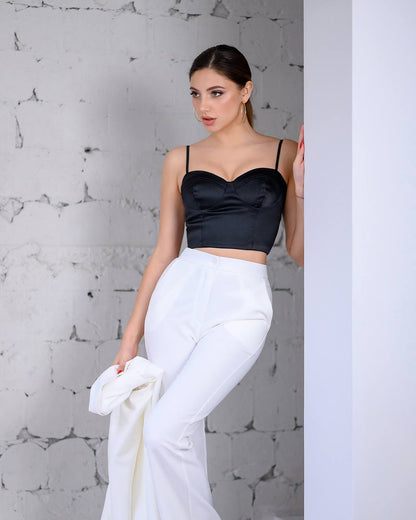 Woman wearing a black satin top and white pants against a textured gray wall.