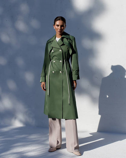 Person wearing a green trench coat standing against a white wall with shadows.