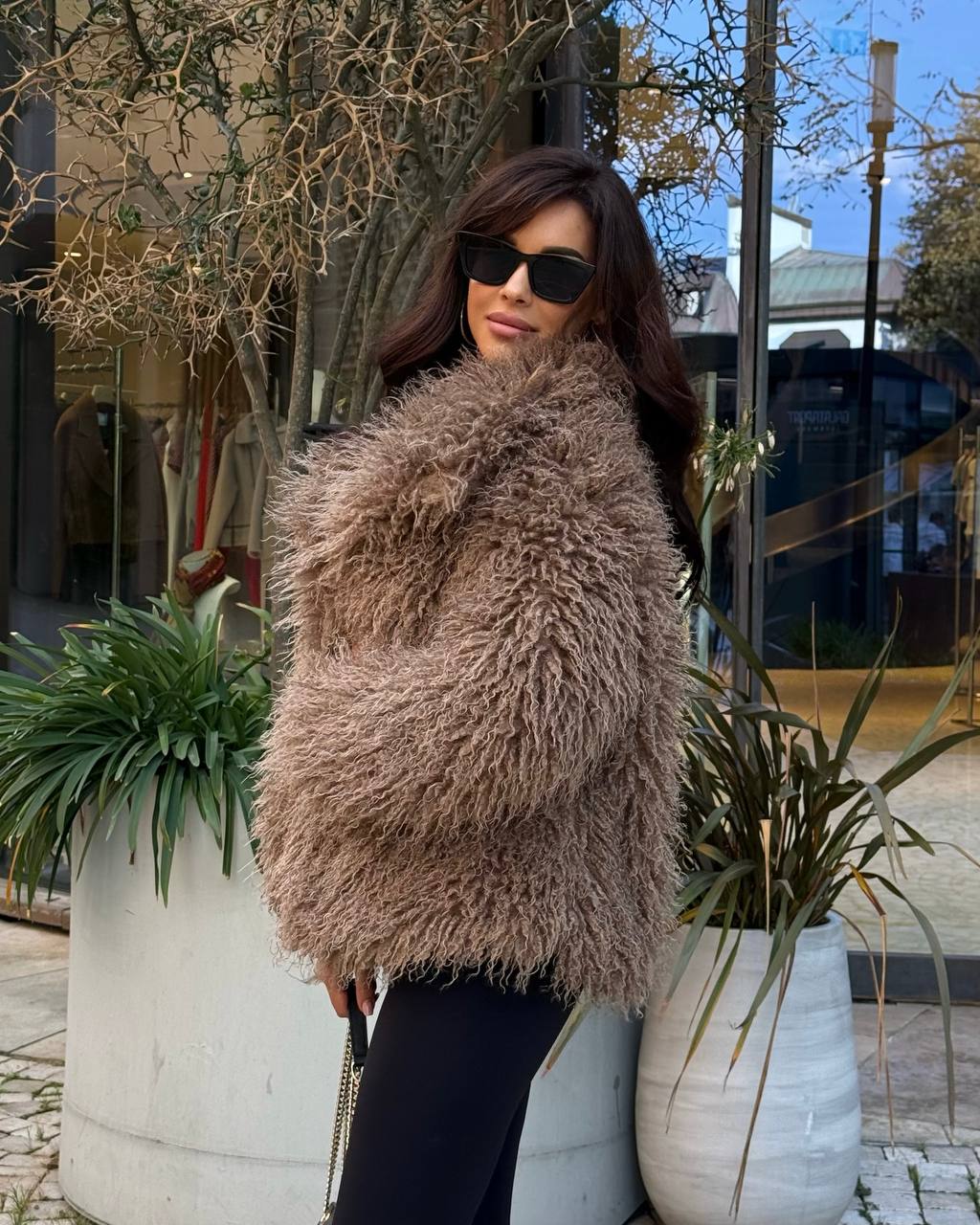 Woman wearing a fluffy brown vest standing outdoors with plants and a building in the background