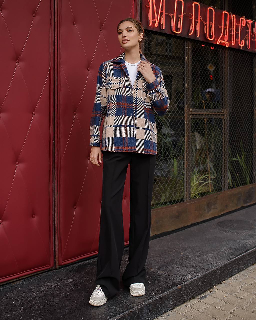Person wearing a plaid shirt and black pants standing in front of a red wall with a neon sign.